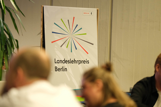 Unscharf sind Menschen vor einem Banner zu sehen, auf dem "Landeslehrpreis Berlin" steht