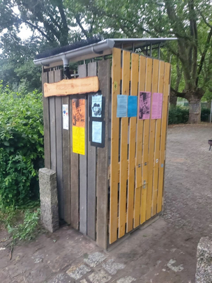 Fertige Regentonne am Parkcafé Auf dem Foto ist die fertige Regentonne zu sehen. Sie steht auf einer gepflasterten Fläche und hat eine hölzerne Verkleidung und ein Satteldach. Im Hintergrund sind Bäume und Sträucher zu sehen.