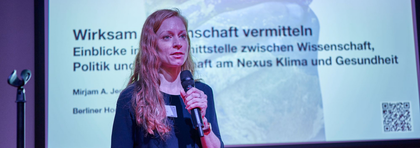 Dr. Mirjam Jenny hält ein Mikrofon in der Hand. Hinter ihr an der Wand ist zu lesen: „Wirksam Wissenschaft vermitteln. Einblicke in die Schnittstelle zwischen Wissenschaft, Politik und Gesellschaft am Nexus Klima und Gesundheit“