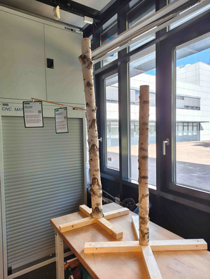 Infobaum Testaufbau in der Glasbox Das Foto zeigt den Testaufbau von zwei selbstgebauten Infobäumen aus Birkenholzstämmen und hölzernen Füßen. Die Bäume stehen in einem Raum auf einer Werkbank vor einem Fenster.