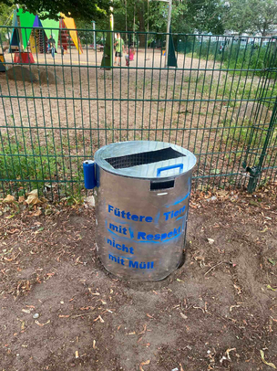 Der tiersichere Mülleimer im Einsatz auf dem Sparrplatz im Sprengelkiez Auf einem erdigen Untergrund und vor einem Zaun steht ein mit Alublech verkleideter Mülleimer, der oben einen Deckel aus dem gleichen Metall hat mit einer großen rechteckigen Öffnung und einem Griff am Deckel. In blauer Schrift steht auf dem Mülleimer "Füttere Tiere mit Respekt, nicht mit Müll"