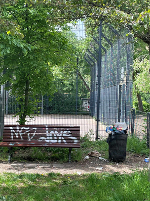 Müll im öffentlichen Raum - Sprengelkiez Wedding In einem Park steht eine mit Graffiti besprühte Parkbank. Daneben steht ein runder überfüllter Mülleimer, um den herum weiterer Müll auf dem Boden verteilt liegt. Hinter der Bank befinden sich ein Sportplatz mit hohen Zäunen und ein paar Bäume im Hintergrund