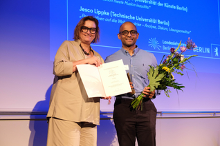 Ina Czyborra und Tharusan Thevathasan posieren auf der Bühne mit Urkunde und Blumenstrauß