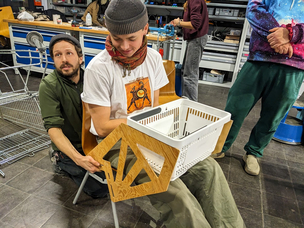 Einblick in den HACKathon: Mock-up der Einkaufskorb-Halterung an einem Stuhl Ein Mann sitzt auf einem Stuhl, der einen Rollstuhlsymbolisieren soll. Ein zweiter Mann hält zwei Adapter aus Sperrholz seitlich an den Stuhl. Vorne ist ein Original-Einkaufskorb eingehängt.