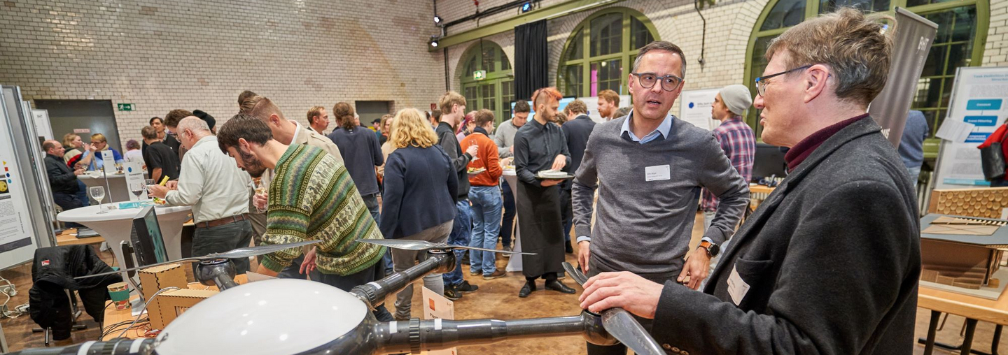 Blick in den Ausstellungsraum: Menschen stehen an Hochtischen und vor Postern. Im Vordergrund: Zwei Personen und eine Drohne.