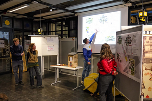 Team "Grünstreifen“ bei der Präsentation: rechts zu sehen ist ein Poster mit den unterschiedlichen geplanten Zonen, während die Studierenden etwas auf der Beamerleinwand zeigen.