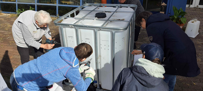 Fünf Personen unterschiedlichen Alters stehen auf einer gepflasterten Freifläche verteilt um einen 1.000 Liter IBC Container und bohren mit Borhmaschinen Löcher in den Container. Das Foto zeigt den Prozess aus einer Nahaufnahme.