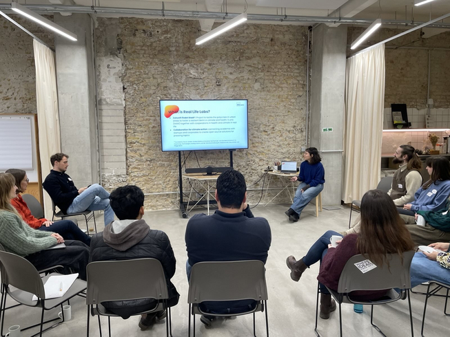 Eine Person sitzt in einem Besprechungsraum des Impact Hubs neben einem Bildschirm und präsentiert eine Folie mit der Aufschrift „What are Real Life Labs“  vor einer kleinen Gruppe von Teilnehmenden, die im Halbkreis auf Stühlen sitzen und zuhören