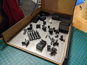 Bastelpackung Miniatur-Supermarkt - die Magnete zur temporären Fixierung der Möbel fehlen noch. Eine Holzkiste wird aufgeklappt. Darin befindet sich eine Metallplatte, auf der kleine 3D-gdruckte Möbel mit Magneten befestigt werden können.