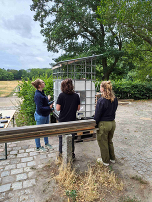 Hackathon im Parkcafé, Bau der Regentonne, Studi (links) baut mit zwei Gästen Das Foto zeigt den Bau einer Regentonne im Parkcafé. Die Regentonne wird von einer Studierenden unter Zuschauenden mit verschiedenem Werkzeug und Material erbaut. Im Hintergrund sind Büsche, eine Wiese und Bäume zu sehen.
