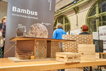 Auf einem Tisch steht ein Modell eines Hauses, das aus Bambus gefertigt ist. Daneben stehen unterschiedliche Konstruktionen aus Bambus: Eine Wabenstruktur, ein kurzes Brett und ein Doppelbrett. Im Hintergrund steht ein Roll-up mit der Aufschrift „Bambus. Das nachhaltige Baumaterial“.