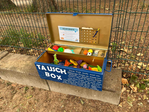 Die Tauschbox im Einsatz auf dem Spielplatz am Sparrplatz im Sprengelkiez Auf einer Sitzbank aus Steinen steht vor einem Zaun eine blau lackierte und geöffnete Kiste, auf der in Weiß "Tauschbox" steht sowie ein kleiner nicht leserlicher Text. In der Kiste befinden sich ein großes und mehrere kleine Fächer, in denen kleinere Kinderspielzeuge - vor allem Sandkastenförmchen - liegen