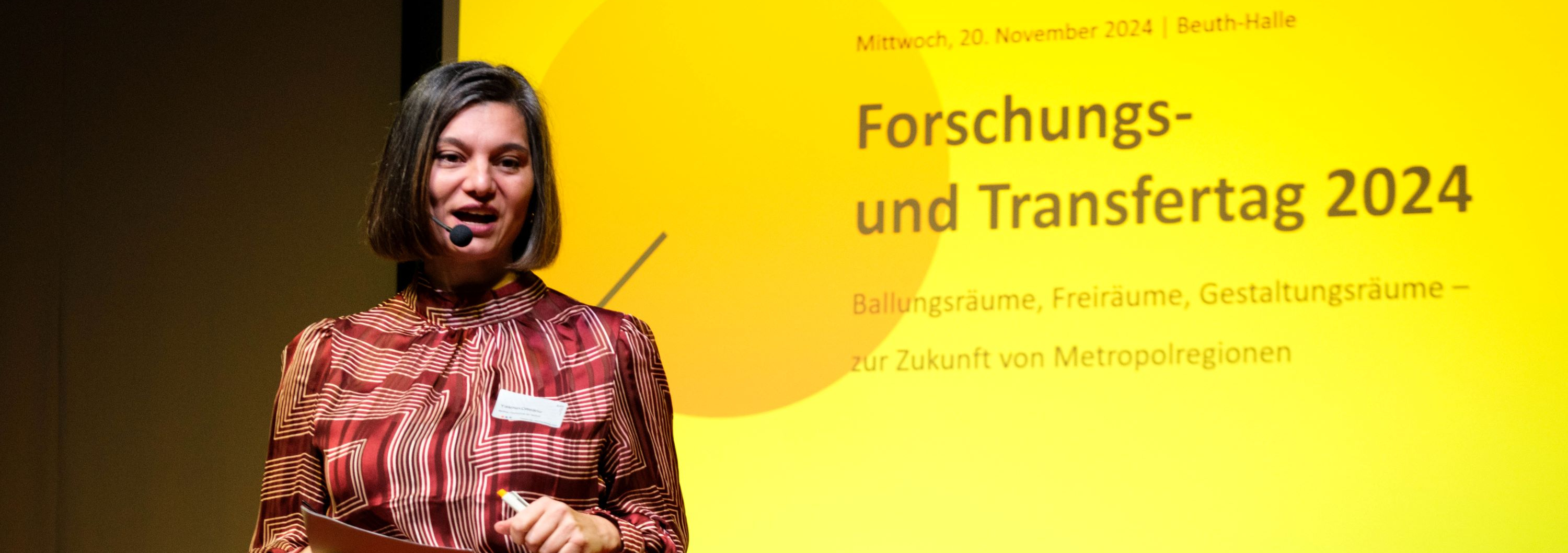 Eine Person spricht auf der Bühne und hält Karten in der Hand. Auf der Leinwand hinter ihr ist zu lesen: Mittwoch, 20. November 2024 | Beuth-Halle Ballungsräume, Forschungs- und Transfertag 2024, Freiräume, Gestaltungsräume – zur Zukunft von Metropolregionen