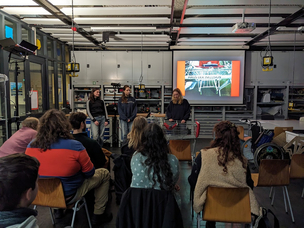 Endpräsentation - Gruppe "Einkaufswagen" präsentiert mehrere Menschen sitzen in der GLASBOX. vorne präsentiert eine Gruppe ihren optimierten Einkaufswagen