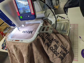 in die Stickmaschine ist ein Rahmen eingespannt, die Stickmaschine stickt erste Buchstaben auf ein großes Stück Stoff