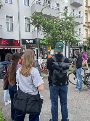 Besichtigung des Weddings mit der Initiative "Sauberer Sprengelkiez" Vor der Kulisse von mehreren mehrgeschossigen Gebäuden und kleineren Bäumen steht eine Gruppe Studierender, die sich auf einer Führung von der Initiative "Sauberer Sprengelkiez befinden. Eine Person hat ein Fahrrad, die anderen stehen mit dem Rücken zur Kamera in Richtung der Leiterin der Führung, die gerade etwas erläutert. Im EG der Gebäude befinden sich Imbisse und andere Dienstleistungen, die nicht direkt zu erkennen sind