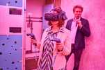 Prof. Dr. Yasmin Olteanu hält zwei Controller in der Hand und trägt eine VR-Brille. Hinter ihr eine Person mit Mikrofon. Der Raum ist in lila Licht getaucht.