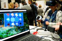 Eine Person trägt eine VR-Brille und Controller. Auf einem Laptopbildschirm im Vordergrund des Fotos ist eine grüne Wiese und eine Wand mit Farbflecken zu sehen.