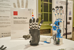 Auf einem Tisch stehen zwei Roboter-Hände: Eine ist grau mit weißen Gelenken, eine ist weiß mit grauen Fingerkuppen und blauen Gelenken. Daneben ein Schild mit der Aufschrift „SIRo Soft Hand“
