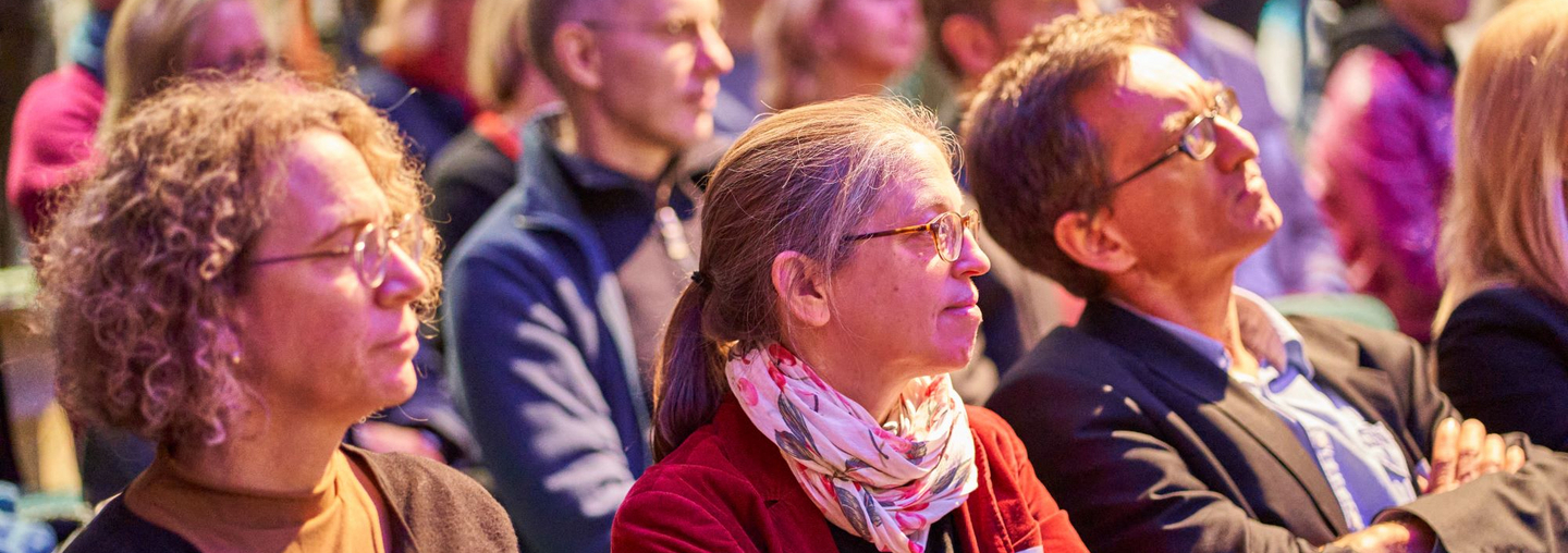 Blick ins Publikum: Vorn im Bild, drei Menschen mit Brille, die ihren Blick nach rechts neigen.