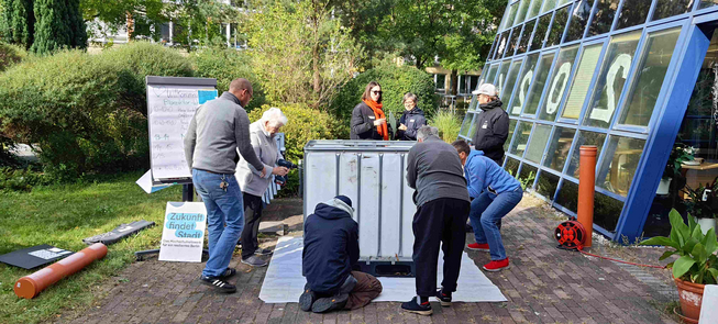 Acht Menschen unterschiedlichen Alters stehen auf einer gepflasterten Freifläche um einen 1.000 Liter IBC Container, aus dem sie den Behälter des Bioreaktors bauen. Vier Personen bohren teilweise stehend, teilweise knieend mit Bohrmaschinen Löcher in den Container. Eine Person links neben dem Container beobachtet die anderen Personen. Drei Person stehen neben dem Container und tauschen sich aus. Links im Bild ist eine Wiese zu sehen, hinten Büsche und rechts die Glasfassade eines Gebäudes. Zudem ist eine Tafel mit den einzelnen Tagesordnungspunkten zu sehen, der Text darauf ist nicht leserlich.