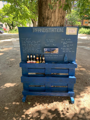 Pfandstation im Einsatz auf dem Sparrplatz im Sprengelkiez Vor einem Baum in einem Park steht eine blau lackierte Pfandstation, die aus ehemaligen Euro-Paletten und einer Holzplatte gebaut wurde. Im unteren Teil stehen Pfandflaschen in dafür vorgesehenen Fächern. Auf dem oberen Teil steht unter der Überschrift "Pfandstation" ein nicht leserlicher Text in Weiß