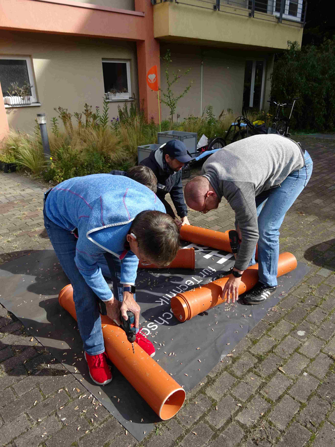Vier Menschen unterschiedlichen Alters bohren auf einer gepflasterten Außenfläche vor einem Gebäude und einer kleinen Grünfläche mit Bohrmaschinen jeweils Löcher in vier orangefarbene Rohre. Zwei Personen stehen dabei über den auf dem Boden liegenden Rohren, zwei sitzen auf dem Boden.