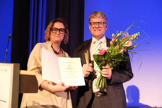 Ina Czyborra und Andreas Modler posieren auf der Bühne mit Urkunde und Blumenstrauß