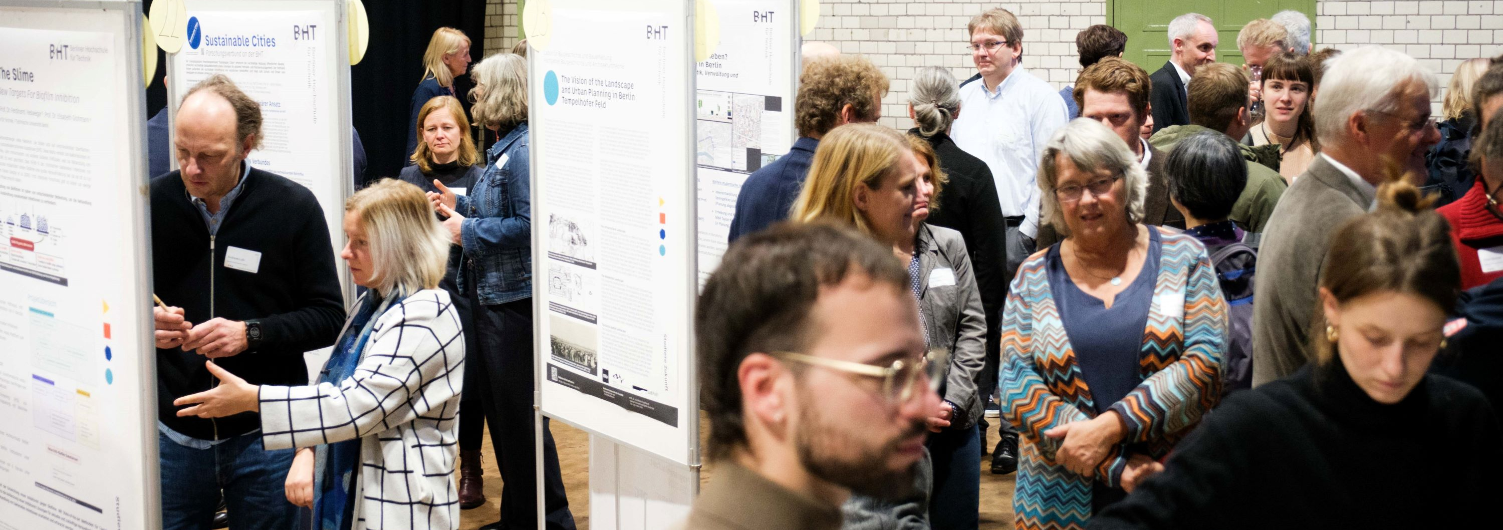 Blick in die Veranstaltungshalle. Viele Menschen stehen in Gruppen und sprechen, dazwischen Stellwände mit Postern