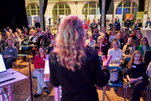 Eine Person mit blonden langen Haaren von hinten, dahinter Sitzreihen mit dem Publikum und der Ausstellungsraum mit Postern.