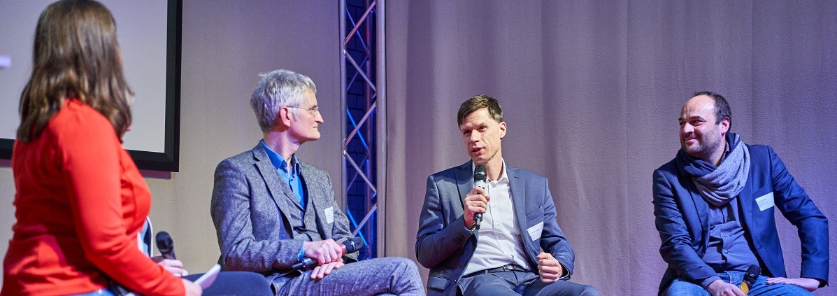 Vier Personen sitzen auf Stühlen auf der Bühne und diskutieren.